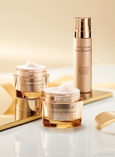 A high-end product photography shot featuring two open cream jars and one serum bottle arranged on a gold reflective surface with a soft, warm beige background.