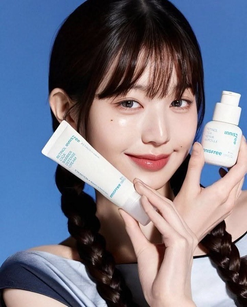 A close-up portrait of a celebrity holding a retinol cream and an ampoule, utilizing a clean, high-trust aesthetic with a solid blue background.