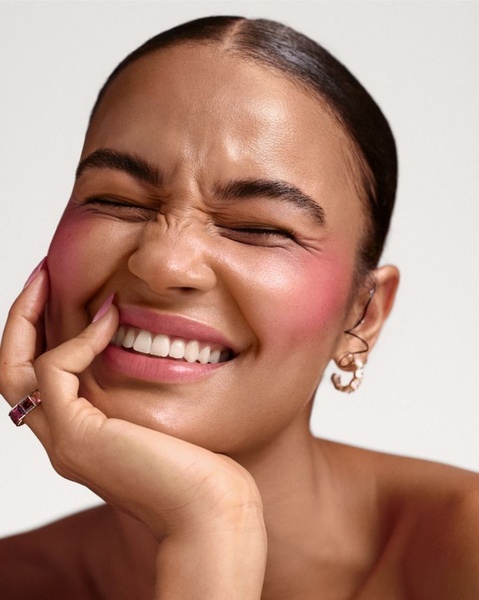 A close-up portrait of a woman applying blush, showcasing the texture and pigment of the product through an expressive, candid reaction.