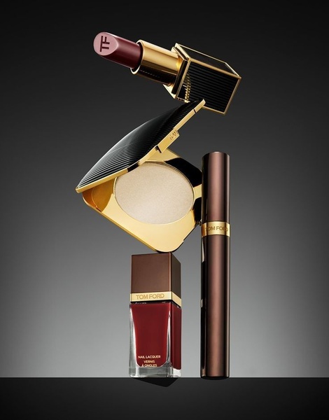 A high-end studio shot featuring a lipstick, powder compact, and nail lacquer balanced in an artistic, minimalist stack against a dark, moody background.