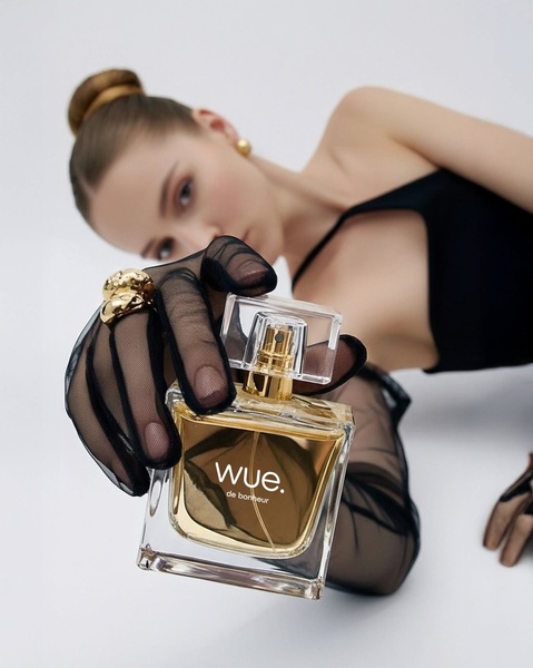 A close-up aesthetic shot of a model in sheer gloves holding a luxury fragrance bottle toward the camera, emphasizing elegance and product detail.