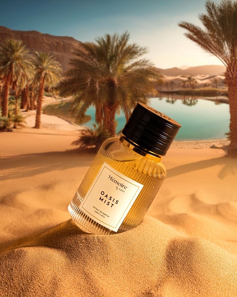 A high-end product shot featuring a Mémoire perfume bottle resting on soft golden desert sand against an oasis backdrop.