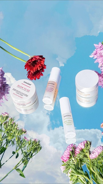 A high-impact skincare hero shot showcasing various serums and creams arranged against a bright blue sky background with floral elements.