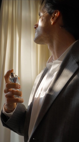 A man in a sharp suit applies fragrance in a sunlit, elegant setting.