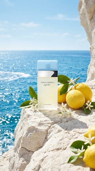 A high-end perfume bottle resting on a sun-drenched cliff against a vibrant blue ocean background.