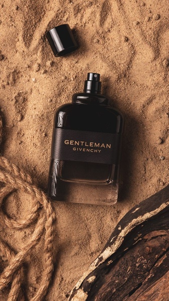 A high-end, top-down product hero shot featuring a perfume bottle resting on natural sand with rustic rope and driftwood textures.