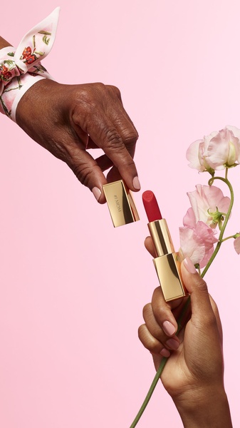 A high-end, close-up lifestyle shot featuring two hands interacting with a gold-cased lipstick, set against a soft pink background with flowers.