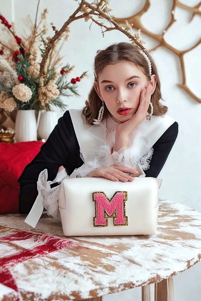 A refined, high-end lifestyle shot featuring a white vanity bag with a prominent pink letter patch, posed next to dried floral decor.