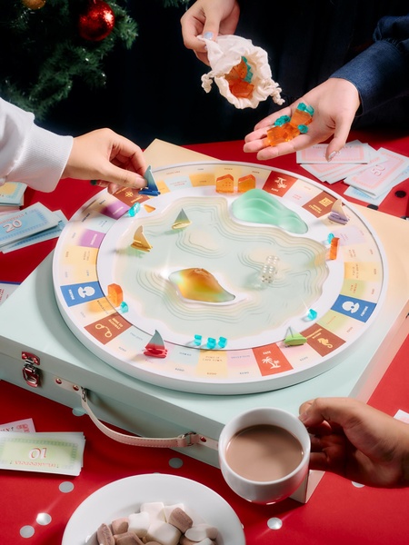 A high-angle tabletop view of a board game in progress, featuring hands interacting with components, tokens, and a cup of coffee.