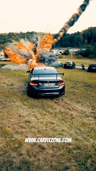 A high-performance BMW 2 Series sits on a grass field while a massive cinematic explosion occurs in the background, creating a high-contrast action scene.