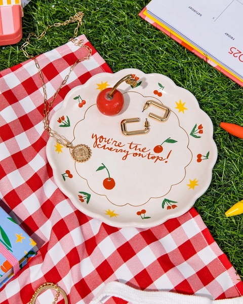 A close-up shot of a decorative cherry-themed ceramic plate, gold necklace, and earrings arranged on a red-and-white checkered picnic blanket.