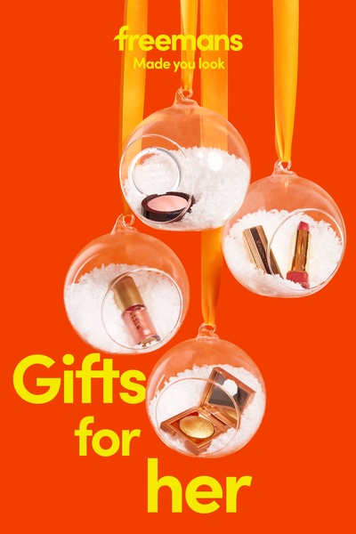 A visually striking product photography shot featuring various makeup items nestled inside glass ornaments against a bright orange background.
