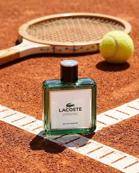 A close-up product shot of a perfume bottle placed on the white lines of a clay tennis court, framed by a wooden racket and ball.