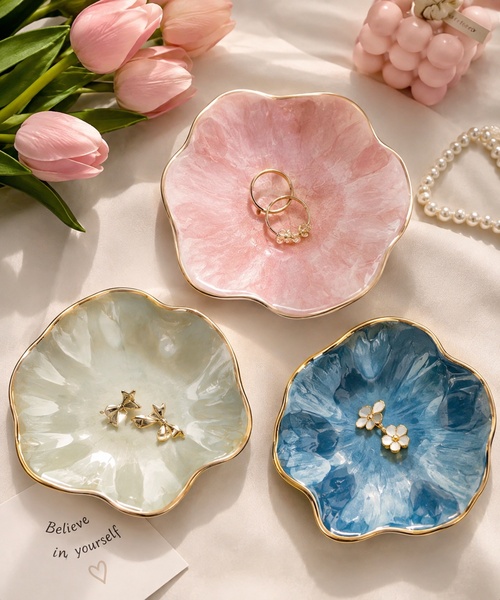 A high-quality lifestyle shot featuring three floral-shaped jewelry trays in pastel colors, arranged with delicate props to evoke a luxury home aesthetic.