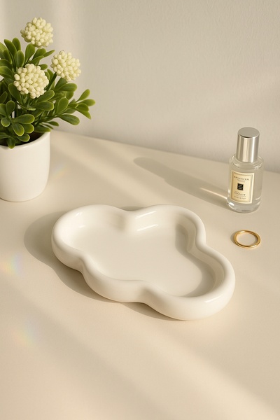 A clean, minimalist product shot featuring a white, cloud-shaped ceramic tray displayed next to a small bottle and a gold ring.