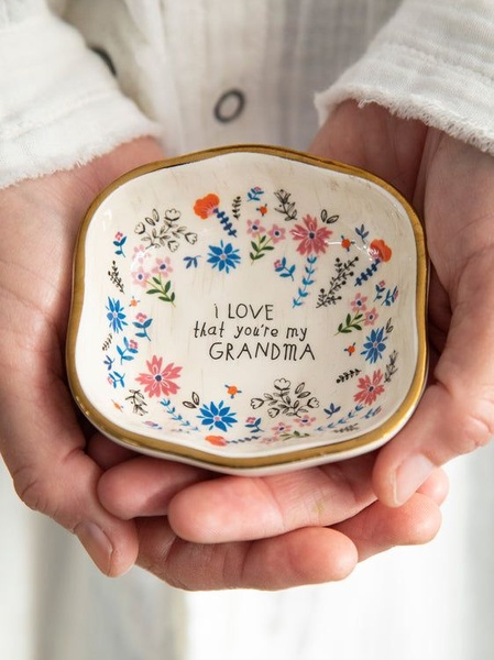 A close-up photograph of a pair of hands holding a small, floral-patterned ceramic dish inscribed with a sentimental message.