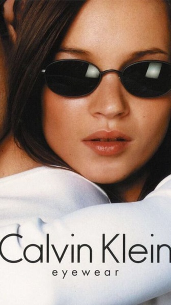 A close-up fashion portrait of a model wearing dark-tinted oval sunglasses, utilizing high-contrast lighting and clean brand typography.