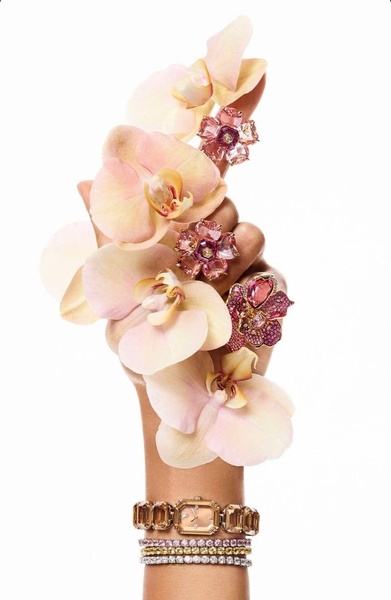 A close-up vertical shot showing a hand wearing a luxury watch and gemstone rings, surrounded by delicate peach and pink orchids.