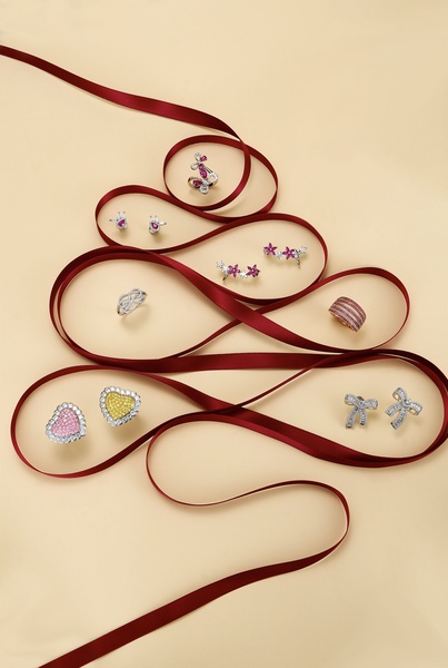 A festive product arrangement featuring various earrings and rings organized along a red silk ribbon shaped into a Christmas tree on a beige background.