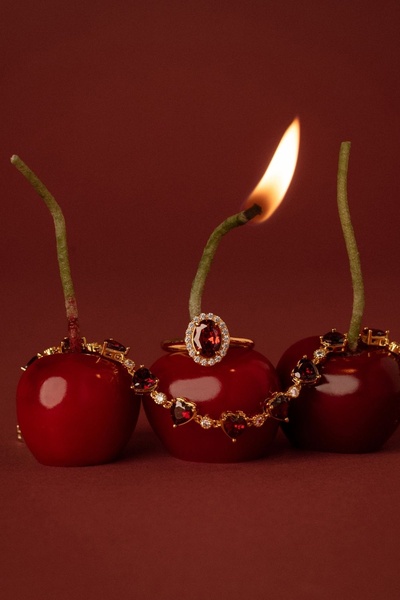 A high-contrast, minimalist jewelry shot featuring necklaces and a ring resting on ripe cherries, with one cherry wick ignited to create a dramatic fire hook.