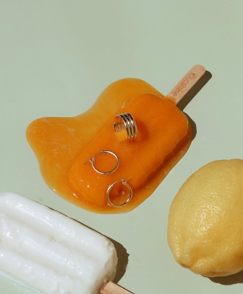A minimalist, high-contrast shot of gold hoop earrings resting on a melting orange popsicle against a pastel background, evoking a fresh summer feeling.