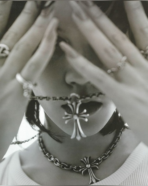 A high-contrast, moody close-up portrait featuring a silver cross necklace, with hands obscuring the model's face to focus on the jewelry.