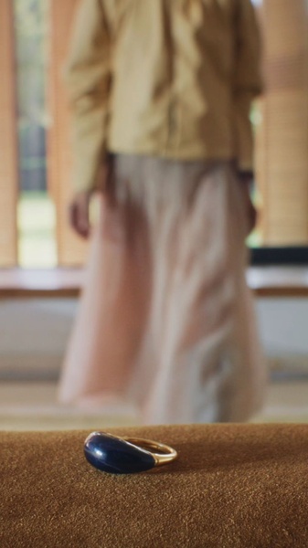 A close-up, soft-focus view of a gold ring with a dark blue stone, placed on a textured tan surface with a blurred person in the background.