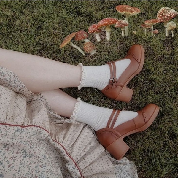 A high-angle, outdoor shot featuring Mary Jane shoes on a grassy field, surrounded by decorative mushrooms to establish a whimsical brand mood.