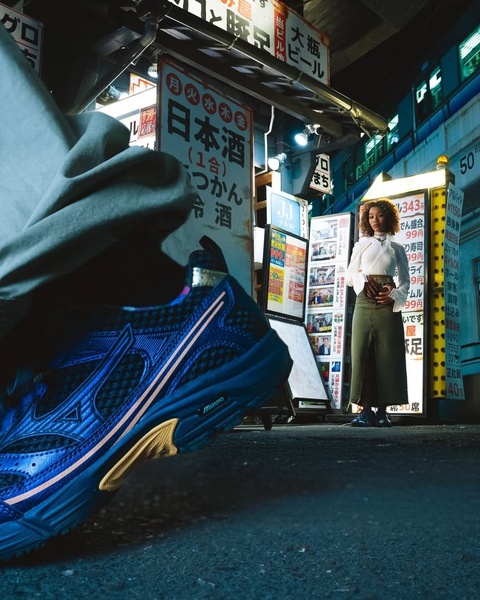 A high-fashion, editorial-style product shot featuring a blue running shoe in the immediate foreground, with a model standing in a blurred, neon-lit Japanese street scene behind.