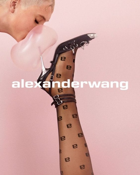 A high-fashion, 4:5 ratio studio portrait featuring a model blowing a bubblegum bubble directly at a uniquely detailed black high heel, utilizing a soft pink background.