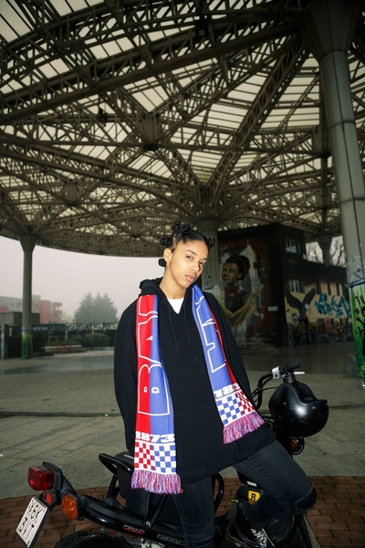 A high-fashion, urban-styled portrait featuring a model wearing a branded scarf in an industrial city setting.