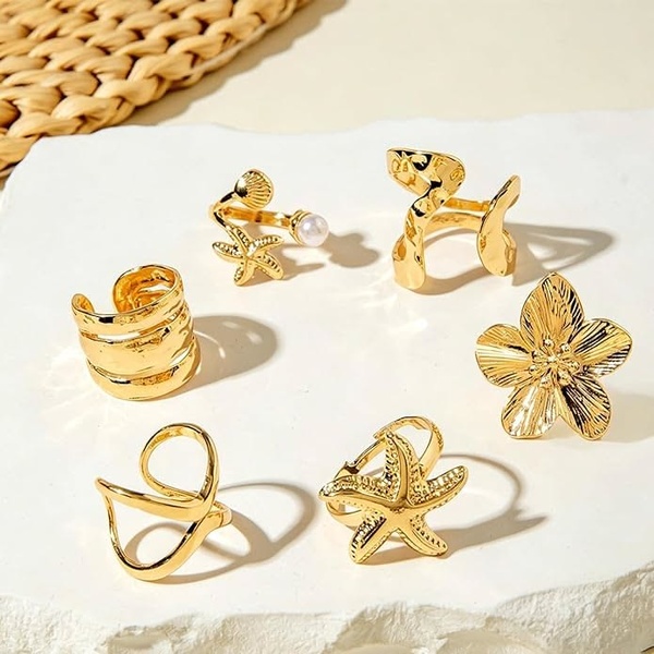 A high-end, clean studio product shot featuring a collection of six gold-toned rings with coastal-inspired designs like starfish and shells.