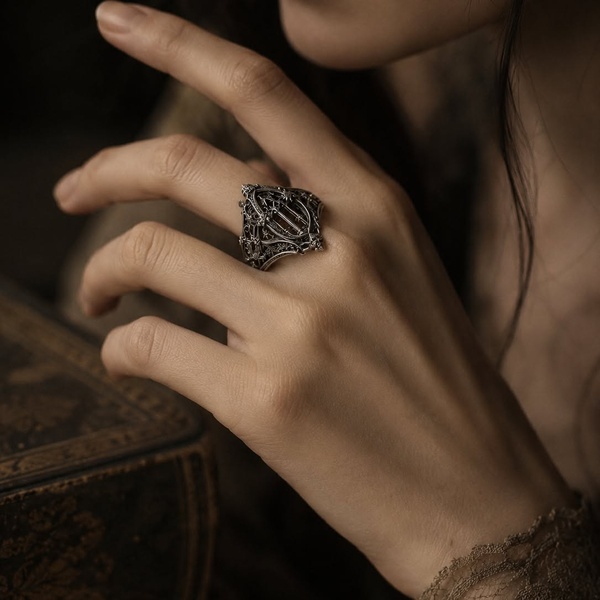 A close-up, moody product shot of a silver gothic filigree ring worn on a hand, emphasizing texture and a dark, refined aesthetic.