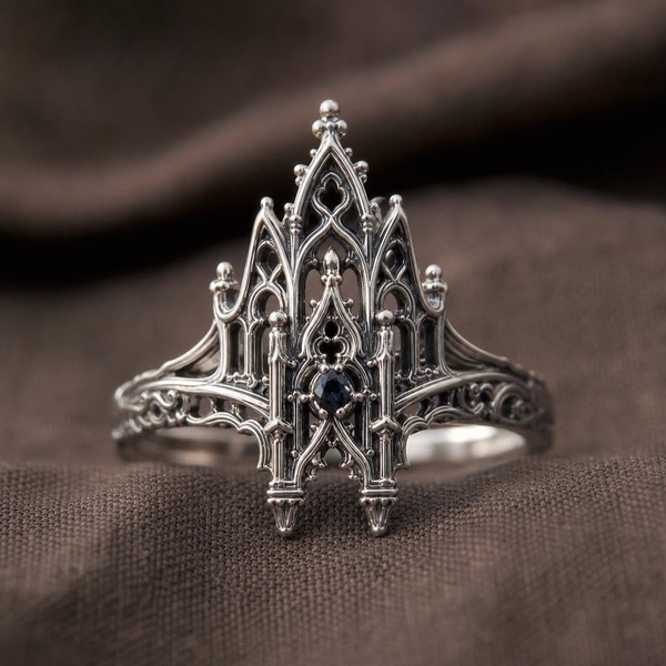 A close-up studio photograph of an intricate silver ring designed with gothic architecture details, set against a dark fabric background.