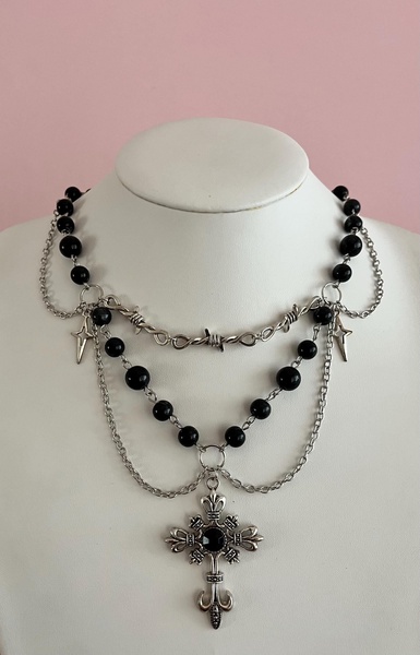 A high-contrast studio shot of a layered gothic-style necklace featuring black beads, metal chains, and a detailed cross pendant on a clean white mannequin.