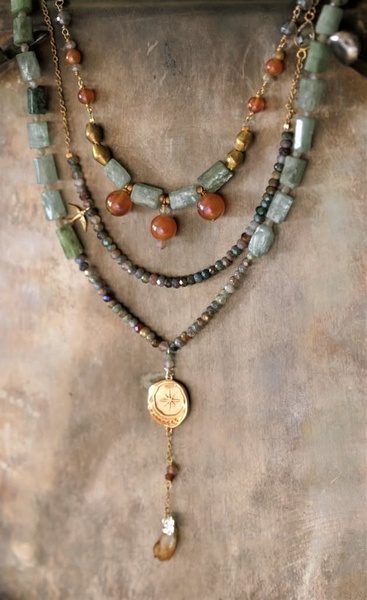 This creative showcases a layered necklace featuring natural stone beads and a gold compass charm against a textured rustic backdrop.