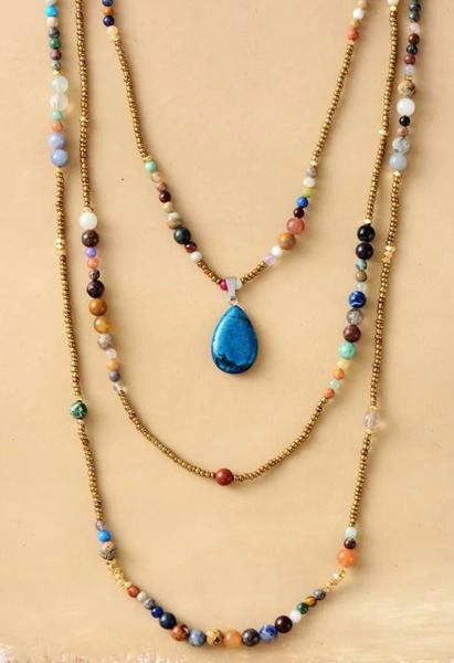 A high-resolution, centered image featuring a triple-strand beaded necklace with a distinct blue pear-shaped pendant on a soft earth-toned background.