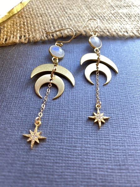 A high-quality product shot featuring gold moon and star earrings against a textured background. The image uses soft, natural lighting to highlight the jewelry's details.
