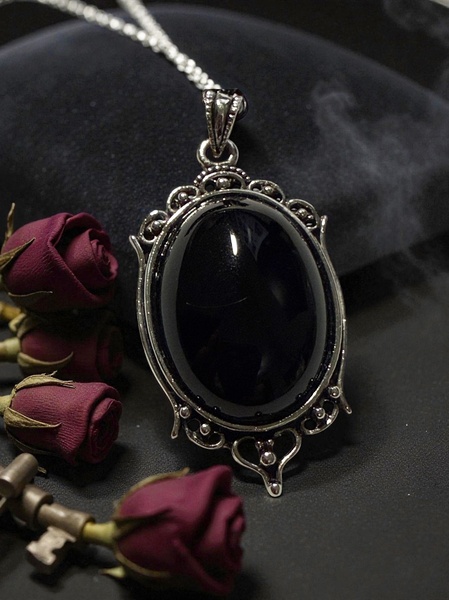 A high-contrast, moody close-up shot of a black stone pendant resting on a dark surface alongside dried roses.