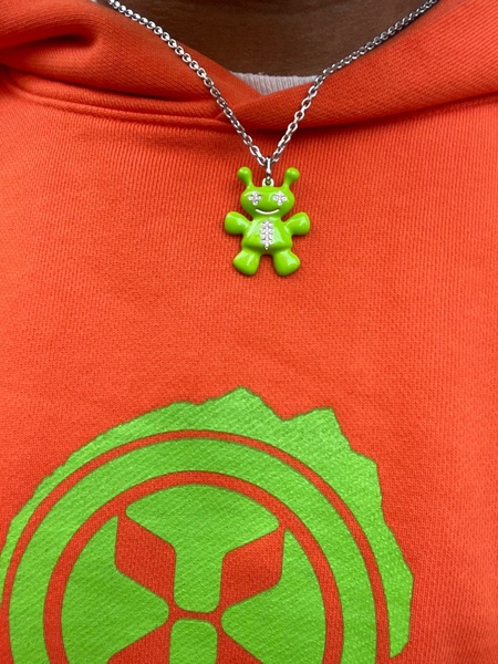 A vibrant green alien pendant necklace is showcased against a matching neon orange hoodie, focusing on the high-contrast aesthetic.