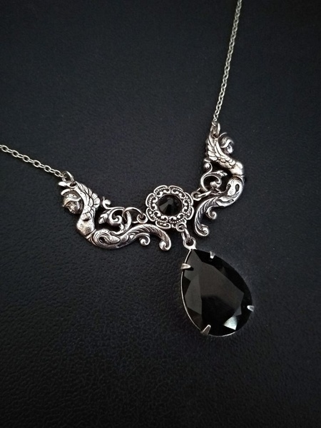 A high-contrast studio shot of a detailed silver necklace featuring gothic motifs and a prominent black teardrop gemstone.