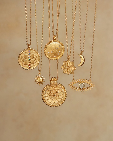 A high-quality studio shot featuring multiple gold necklace pendants floating against a soft, neutral, textured background.