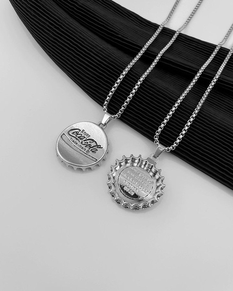 A high-contrast, professional product shot of two bottle cap necklaces arranged against a black pleated fabric background, emphasizing silver metal textures.