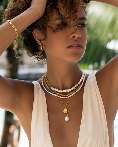A close-up lifestyle image featuring a model wearing multiple gold necklaces with pearl accents, conveying a refined and high-end aesthetic.