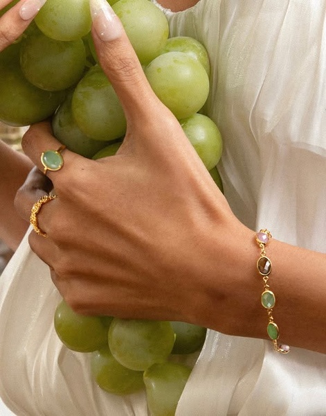 A close-up aesthetic shot of a model wearing a delicate multi-colored gemstone bracelet while holding a bunch of green grapes, emphasizing luxury and lifestyle.