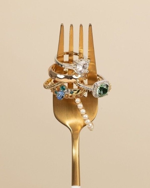 A creative product photography shot featuring a stack of assorted rings resting on a gold fork against a minimalist tan background.
