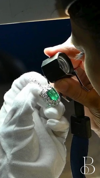 A close-up, first-person view of an expert jeweler inspecting an emerald and diamond ring with a loupe, highlighting craftsmanship and authenticity.