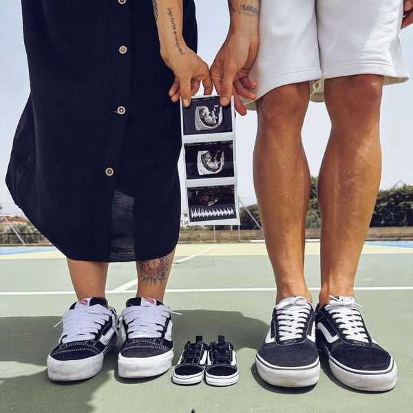 A lifestyle-focused image showing two adults holding ultrasound scans while standing behind a pair of small infant sneakers placed on the ground.