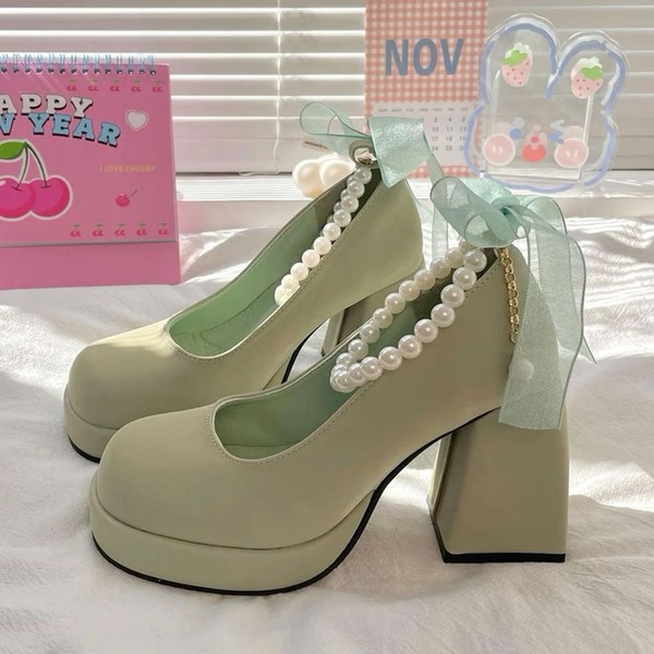 A high-quality 1:1 square product shot of sage green platform heels featuring pearl detailing and ribbon accents in a soft-focus environment.