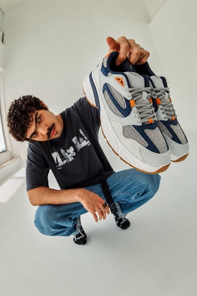A low-angle shot of a person crouching and holding a gray and navy sneaker directly toward the camera, highlighting its texture and design.
